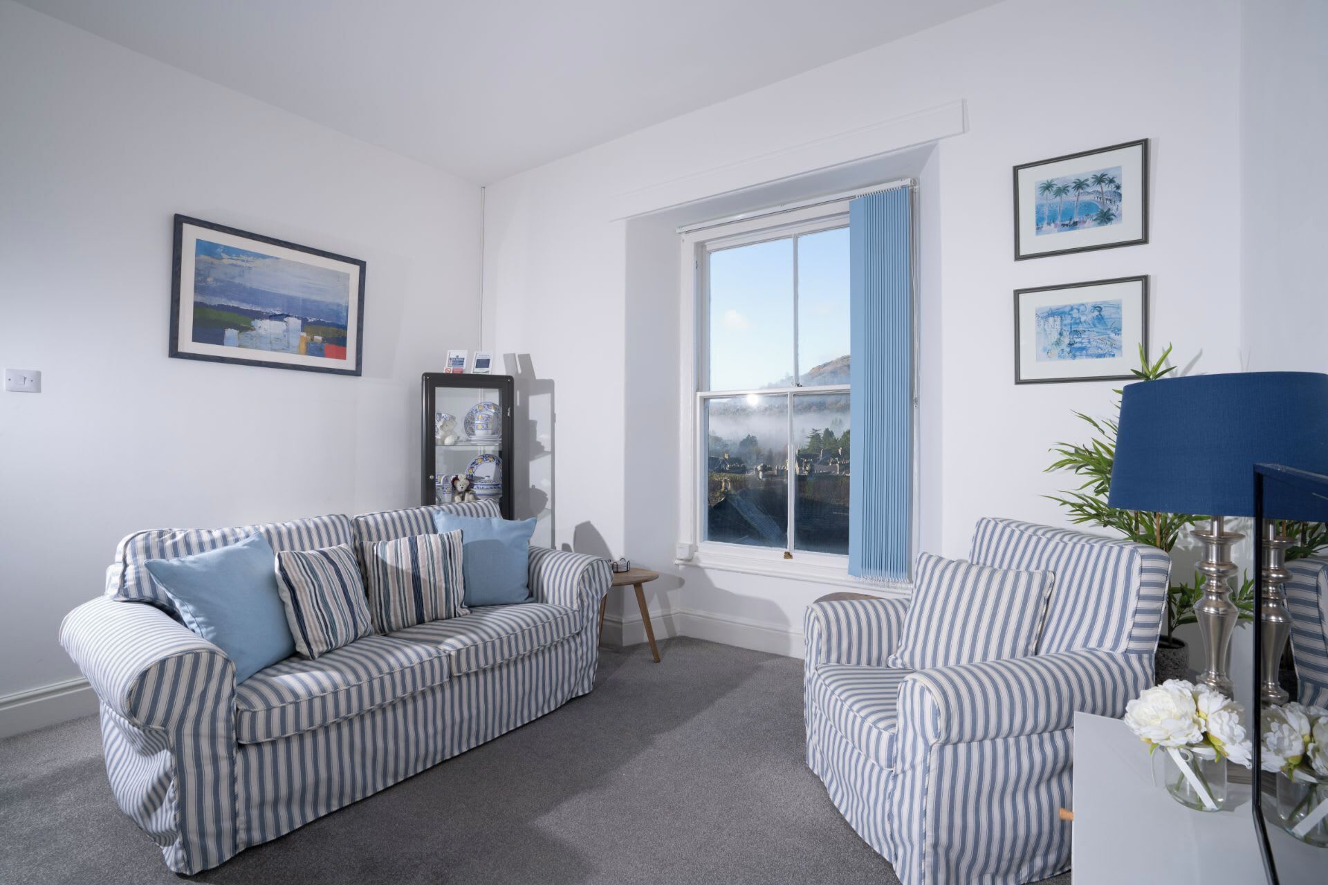 Una sala de estar con un sofá a rayas, un sillón, un aparador con adornos y un ventanal con cortinas.