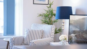 Un fauteuil à rayures, un vase de fleurs blanches, une lampe bleue et un tableau encadré sur une table, dans une pièce dotée d’une fenêtre et d’une plante.