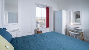 Une chambre avec un grand lit, un bureau avec une chaise, une armoire et une fenêtre donnant sur des immeubles.