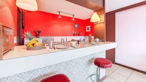 Une cuisine moderne avec des murs rouges, des plans de travail blancs et des appareils en acier inoxydable. On y trouve deux suspensions lumineuses, un tabouret de bar rouge et un pare-feu blanc en carreaux.