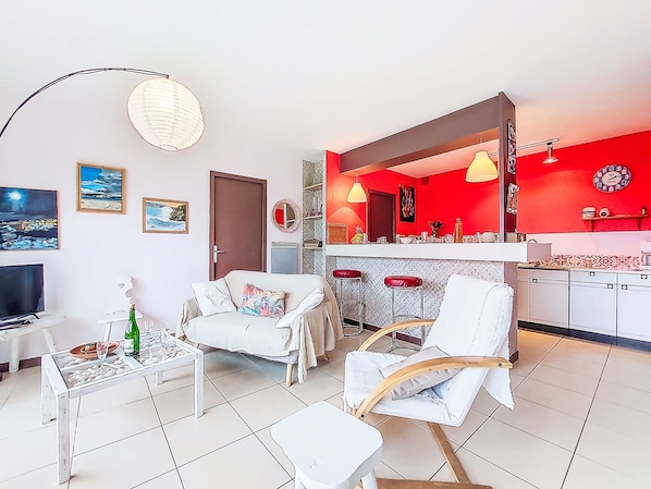 A living room with a sofa, chairs, a coffee table, and a TV. There is a kitchen area with a red wall, a mirror, and bar stools.