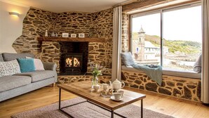 A cozy living room with a stone fireplace, a grey sofa, a wooden coffee table, and a view of a coastal town through a large window. - Emelle - Three Bedroom Cottage, Sleeps 7 (Kingsand)