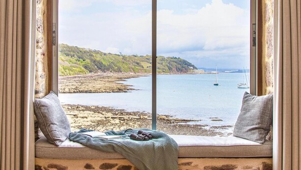 A window seat with a view of a rocky shoreline and calm sea, a sailboat in the distance, and a book and binoculars on the seat. - Emelle - Three Bedroom Cottage, Sleeps 7 (Kingsand)