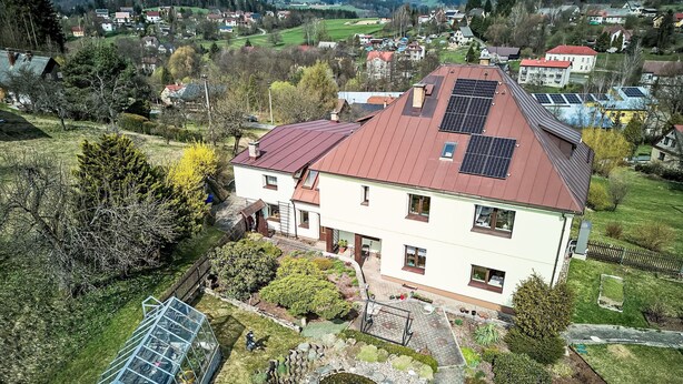 Une maison à deux étages, avec un toit rouge et des panneaux solaires, entourée d’un jardin bien entretenu et d’un petit étang.