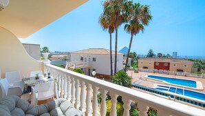 Un balcón con piscina, sillas blancas y vistas a palmeras y edificios.