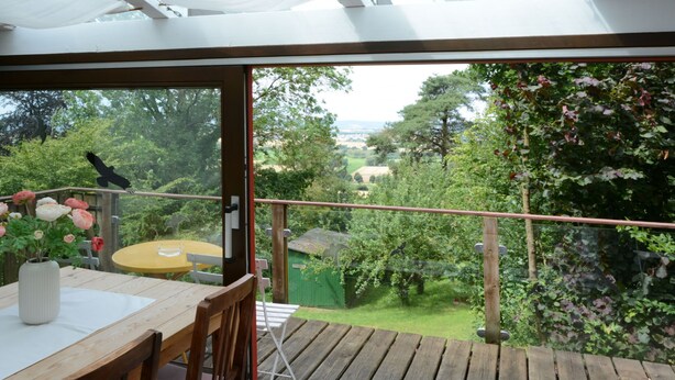 A wooden deck with a table and chairs, a glass door leading to a garden with trees and a view of the countryside. - Vacation home Clobes by Interhome (Wabern)