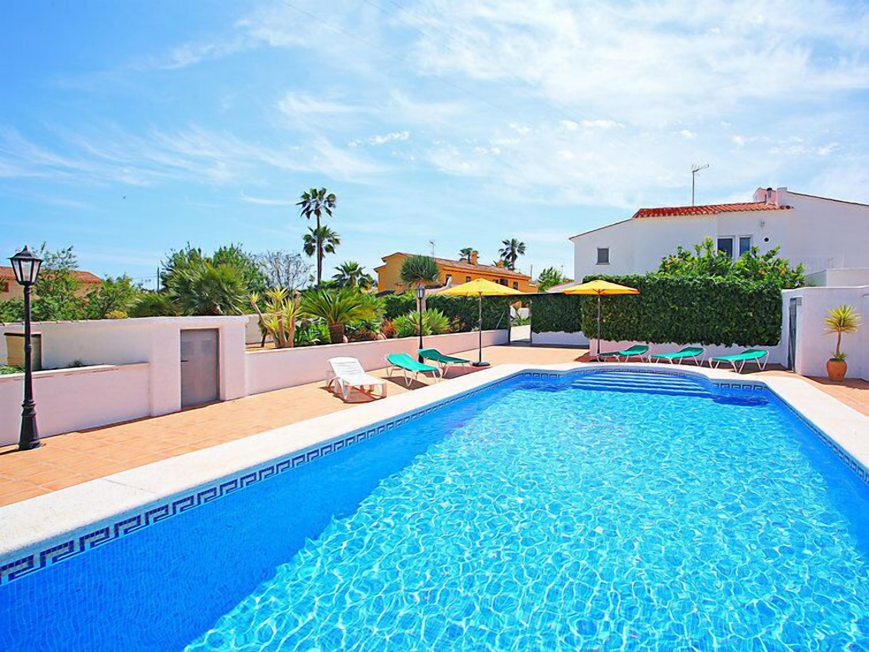 A swimming pool with lounge chairs and umbrellas, surrounded by greenery and buildings.