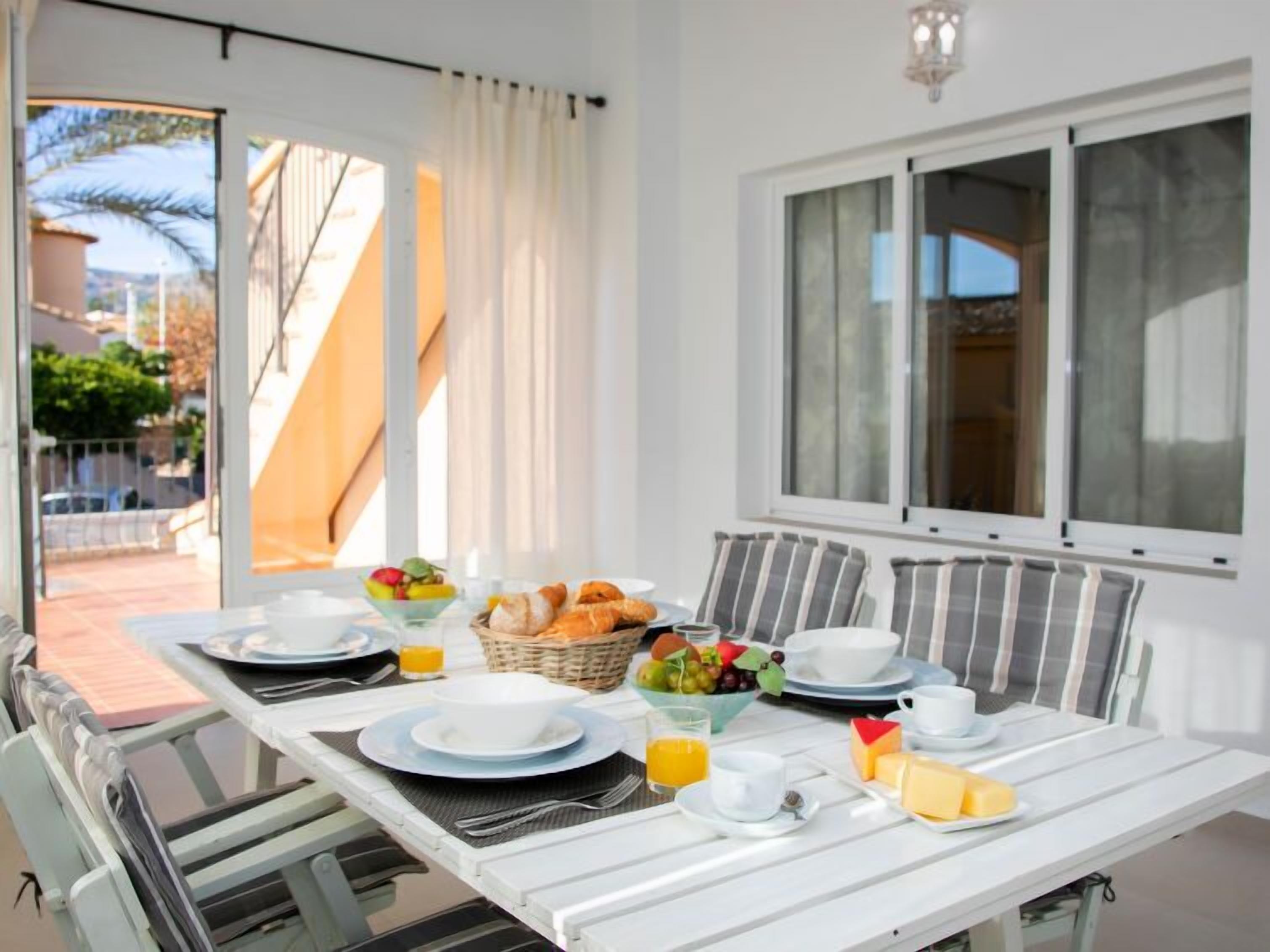 Une table de petit-déjeuner dressée avec des assiettes, des bols et un panier de pain et de fruits.
