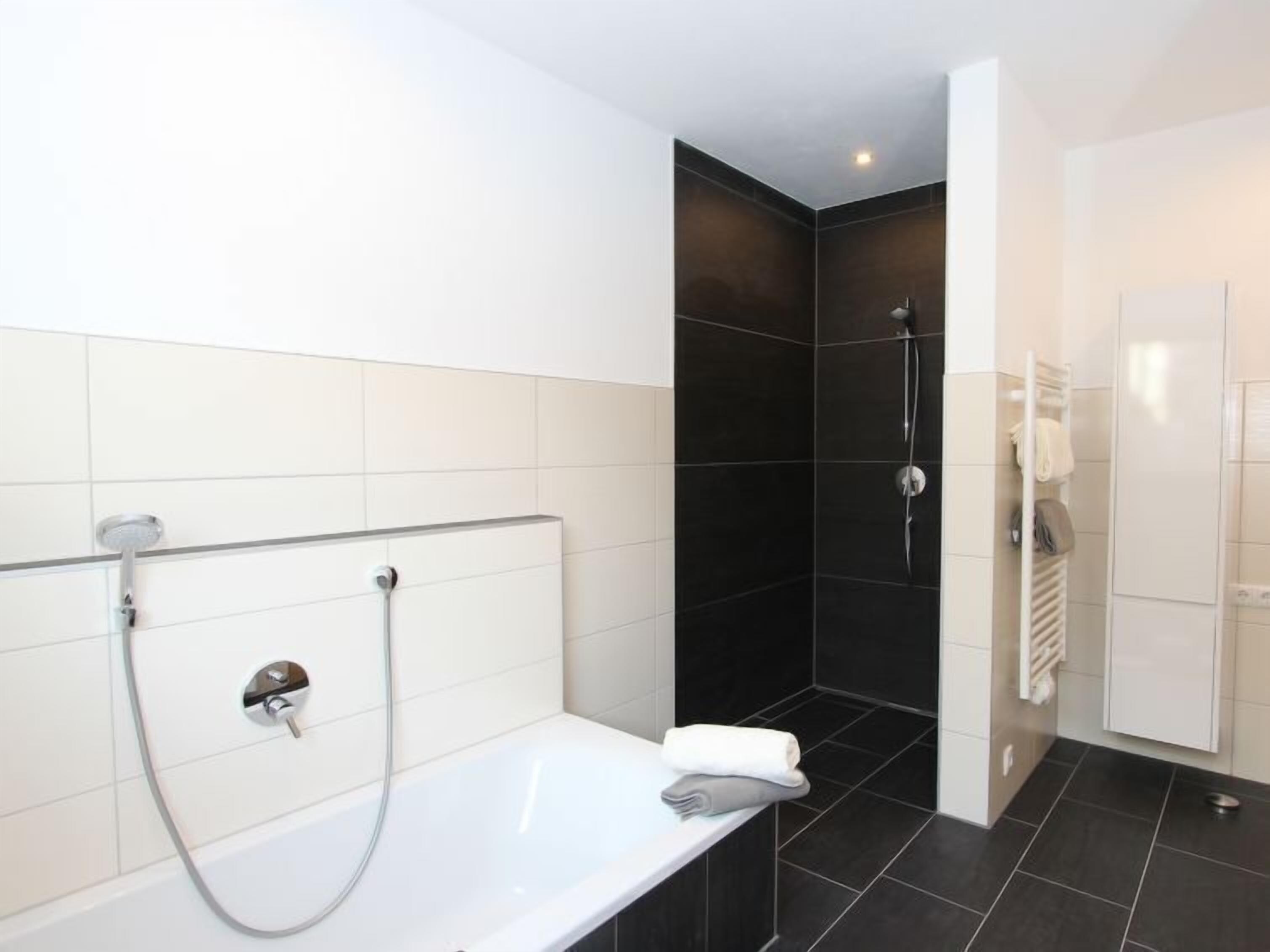 A modern bathroom with a white bathtub, a shower area with dark tiles, and a white towel rack.