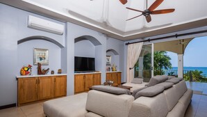 A modern living room with a large sectional sofa, a flat-screen TV, wooden cabinets, and a ceiling fan. - Potrero Beachfront Villa with Pool & Private Chef - Casa Royal (Potrero)