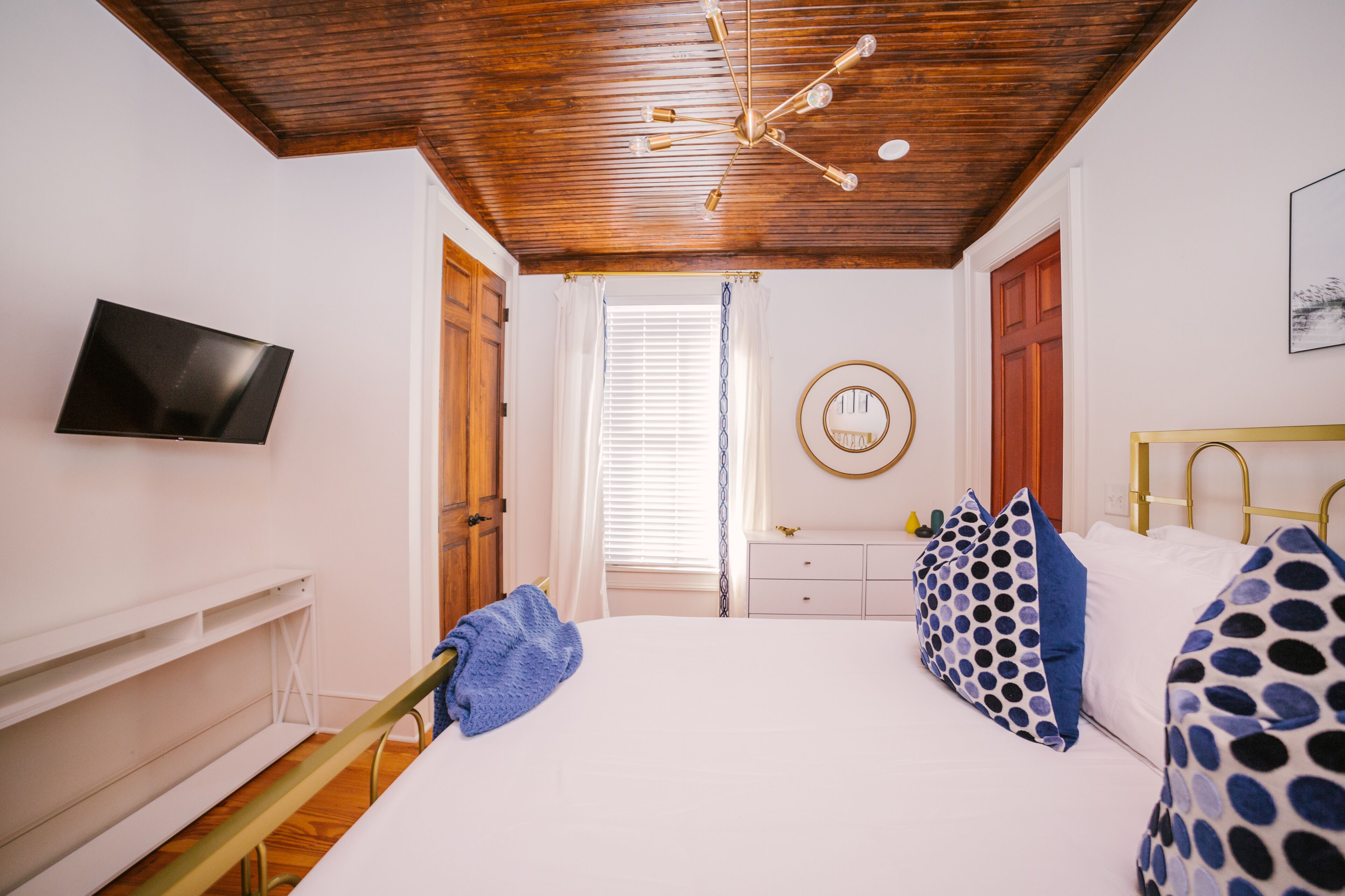 A bedroom with a wooden ceiling, a bed with white bedding, a TV on the wall, and a desk with a chair.
