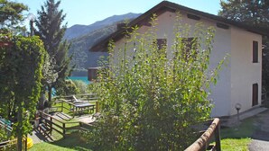 Un bâtiment blanc avec un toit en pente, une terrasse en bois avec une table et des chaises, et une vue sur des montagnes et un lac en arrière-plan.