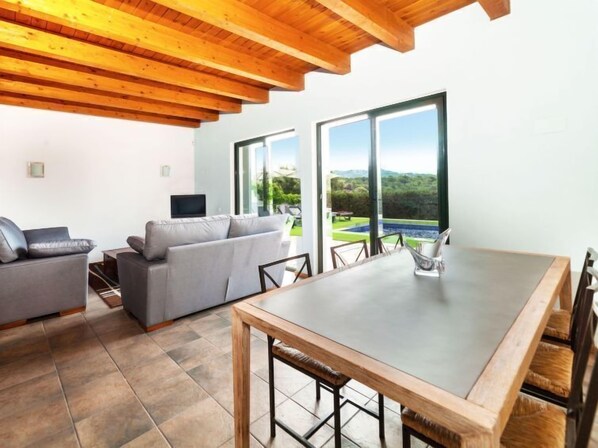 A modern living room with a grey sofa, a wooden dining table, and a TV on a white stand. Large windows offer a view of a garden and a swimming pool.