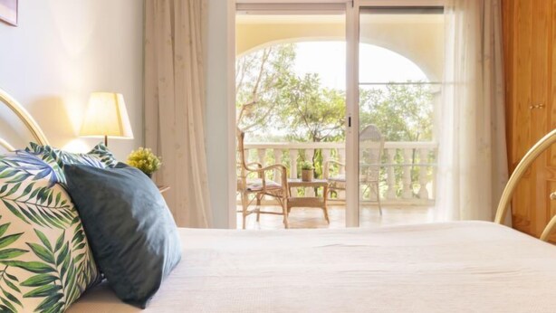 A bedroom with a bed, a nightstand with a lamp, and a view of an outdoor balcony with chairs and a table.