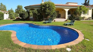 A swimming pool with a curved edge, surrounded by a grassy lawn and a house with a tiled roof and columns.