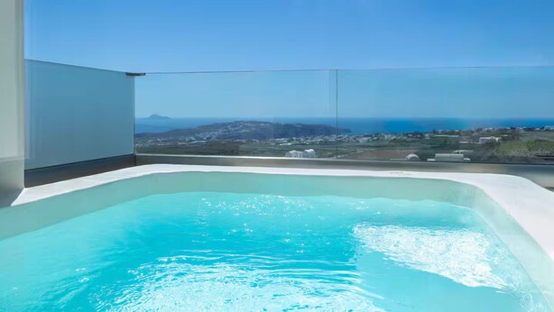 Une piscine à débordement moderne, avec une eau d’un bleu cristallin, entourée d’une balustrade en verre, offrant une vue panoramique sur un paysage côtier et des montagnes lointaines.