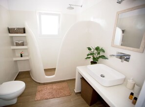 A modern bathroom with a white bathtub, a white sink, and a toilet. There is a small window above the bathtub, a shelf with decorative items, and a potted plant on the sink counter.