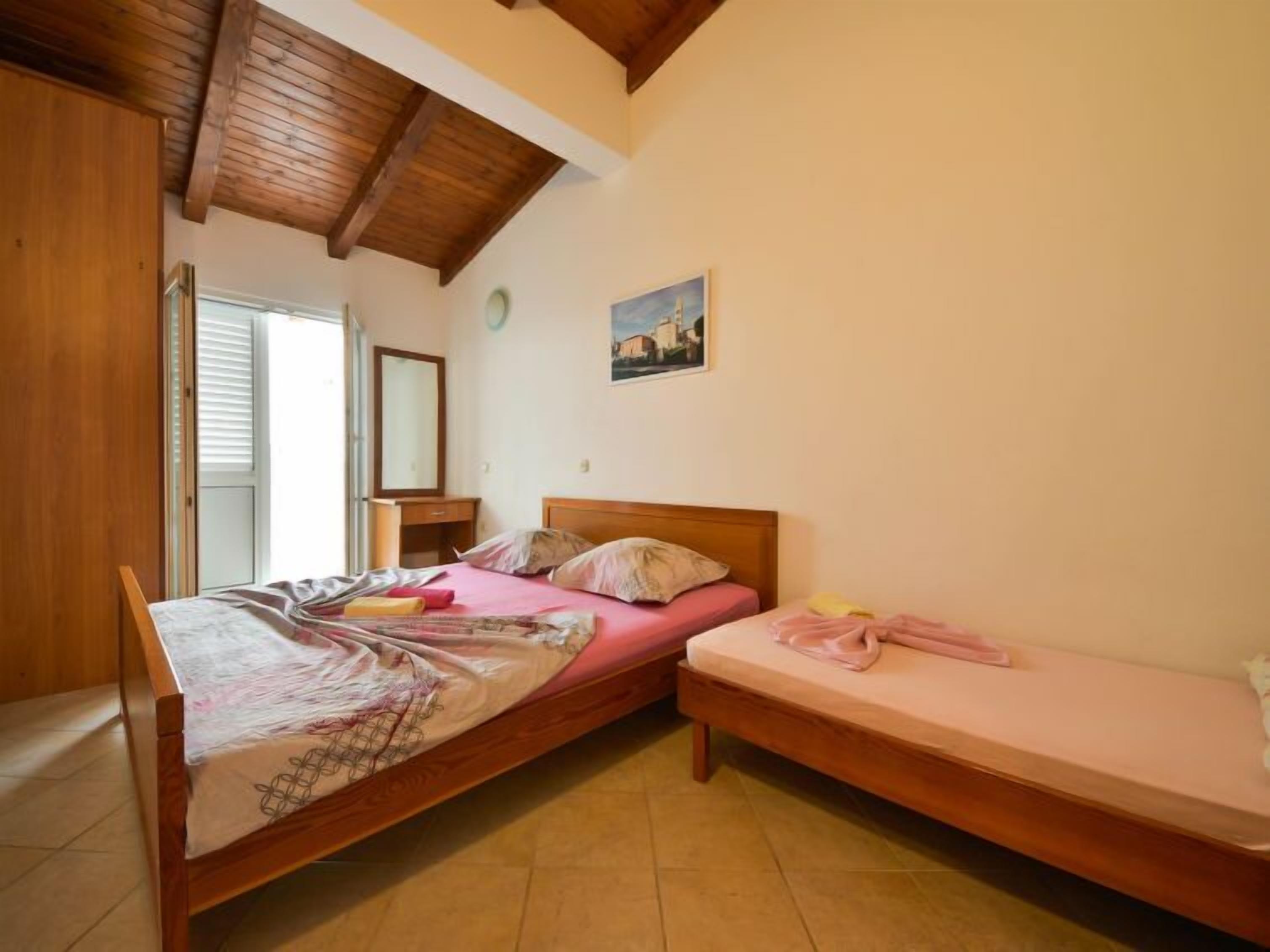 Une chambre avec deux lits, des poutres apparentes en bois au plafond, une fenêtre avec des stores, une table de chevet et un miroir fixé au mur.