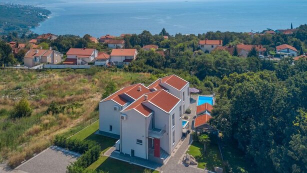 Une maison moderne  à deux étages, avec un toit rouge, entourée d’une végétation luxuriante et offrant une vue sur la mer.