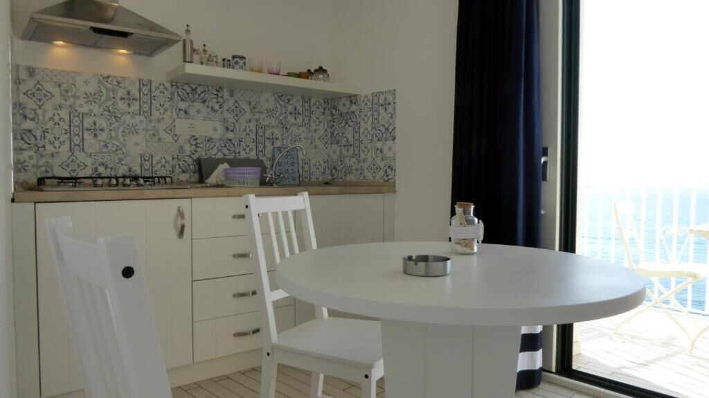 A kitchen with white cabinets, a round table, and a chair. There is a window with curtains and a view of the outside.