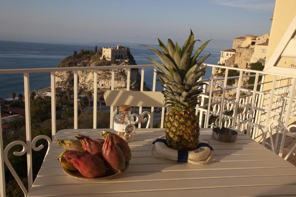 Een balkon met een eettafel, versierd met een ananas en twee rode vruchten, met uitzicht op de kustlijn en een kasteelachtige constructie.