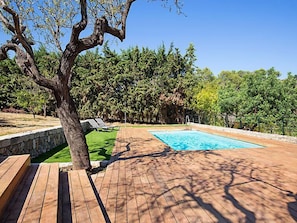 A wooden deck with a swimming pool, surrounded by lush greenery and a stone wall.