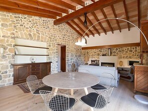 A rustic interior with a stone wall, wooden beams, a round table with chairs, and a kitchen area.