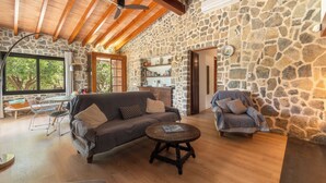 A spacious living area with a stone wall, wooden ceiling beams, a sofa, a round coffee table, and a stone fireplace. - Can Coll by Interhome (Selva)