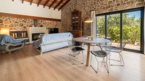 A cozy living room with a stone wall, a fireplace, a sofa, a dining table with chairs, and a large window with a view of trees. - Can Coll by Interhome (Selva)