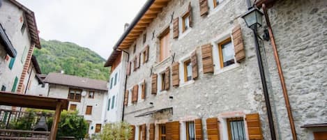 Un edificio de piedra con contraventanas de madera, un balcón, un banco de piedra y una pequeña mesa en un patio.
