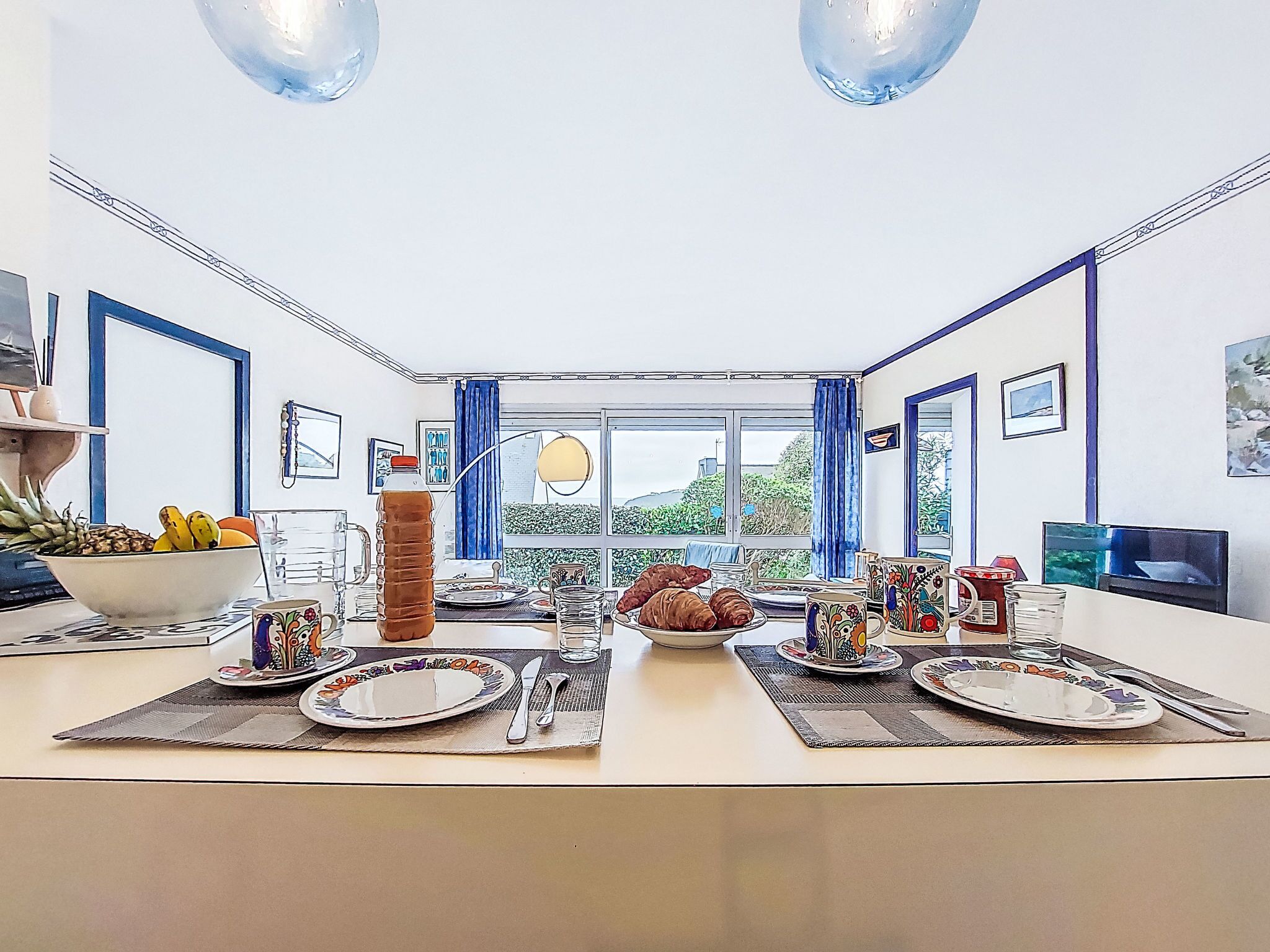 A dining area with a table set for four, a large window with curtains, and a view of greenery outside.