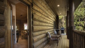 Una cabaña de troncos con una terraza de madera, un comedor con sillas y vistas al bosque circundante.