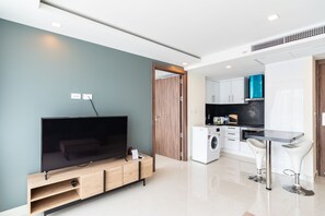 Un salon moderne doté d’un téléviseur à écran plat, d’un meuble de rangement en bois et d’un coin cuisine comprenant un lave-linge et des tabourets de bar.