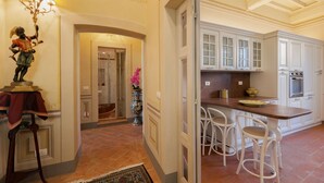 A kitchen with white cabinets, a marble countertop, and a built-in oven. There are two white bar stools and a vase with pink flowers. - Appartamento Zefferini (Cortona)