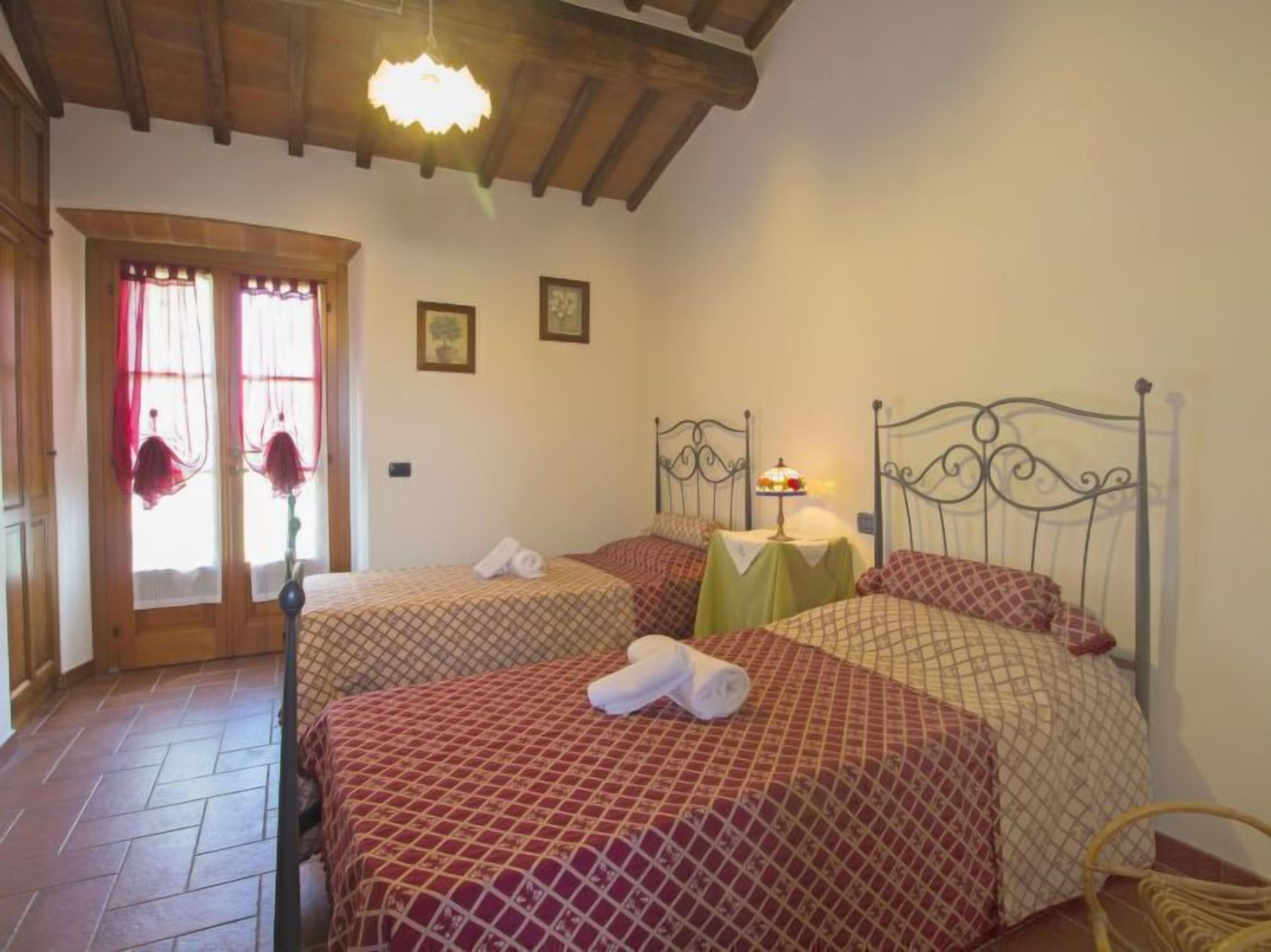 A room with two beds, each with a patterned bedspread, a small table with a lamp, and a window with curtains.