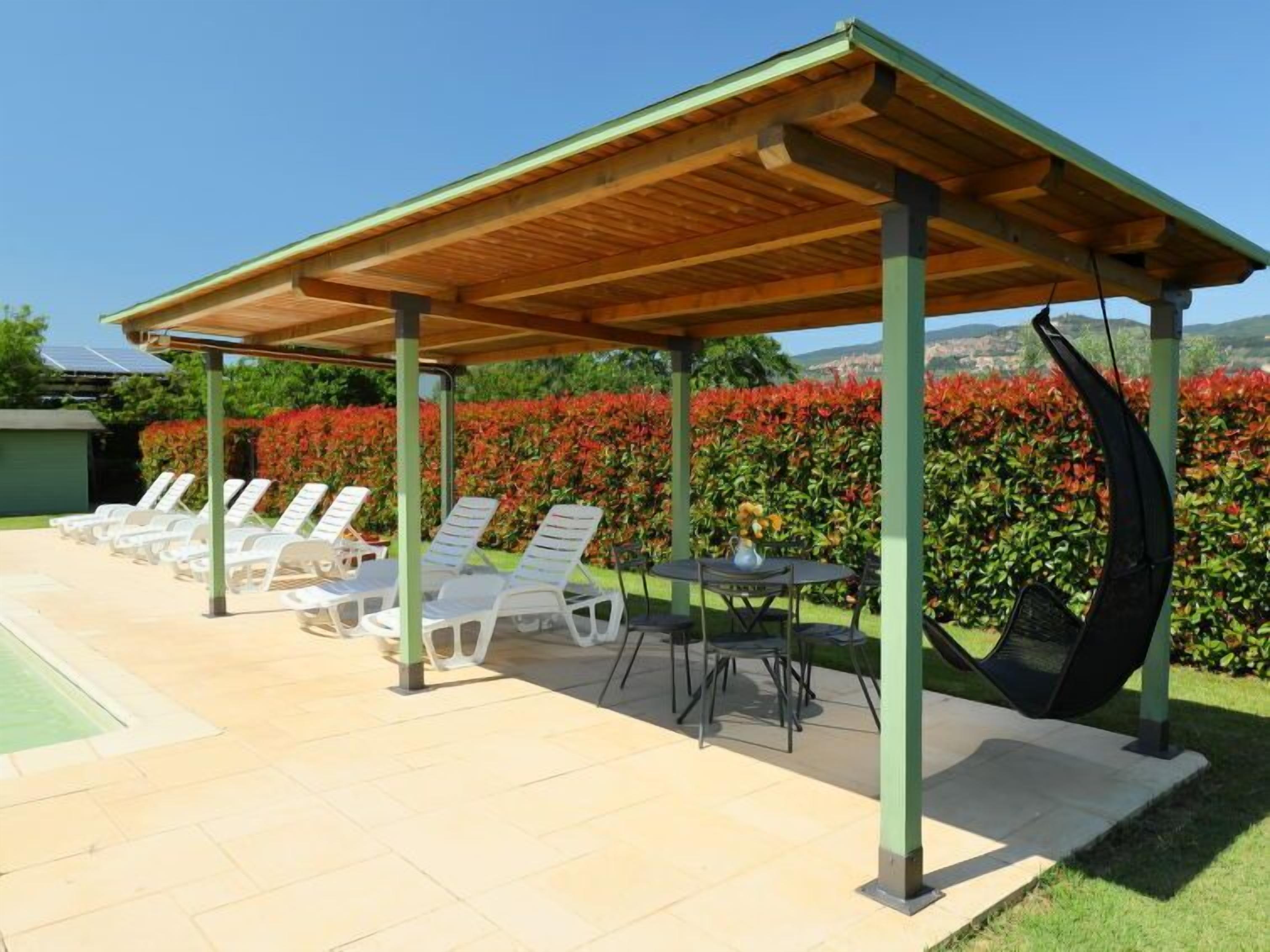 Un espace extérieur aménagé, abrité et doté de fauteuils et d’une table, entouré d’un jardin où poussent des fleurs rouges.