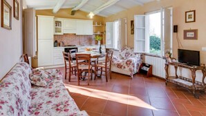 A spacious living room with a sofa, a dining table with chairs, a TV, and a kitchen area with cabinets and a window. - Il Fienile del Colle by Interhome (Pescia)