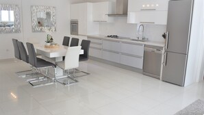 A modern kitchen with white cabinetry, a stainless steel refrigerator, and a dining area with a white table and gray chairs.