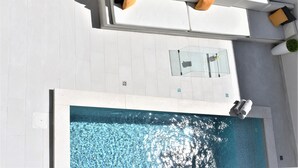 A rooftop pool area with a glass table, white lounge chairs, and a small seating area with cushions.