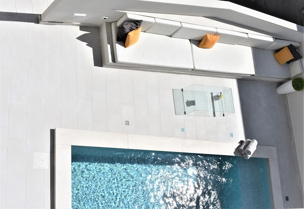 A rooftop pool area with a glass table, white lounge chairs, and a small seating area with cushions.