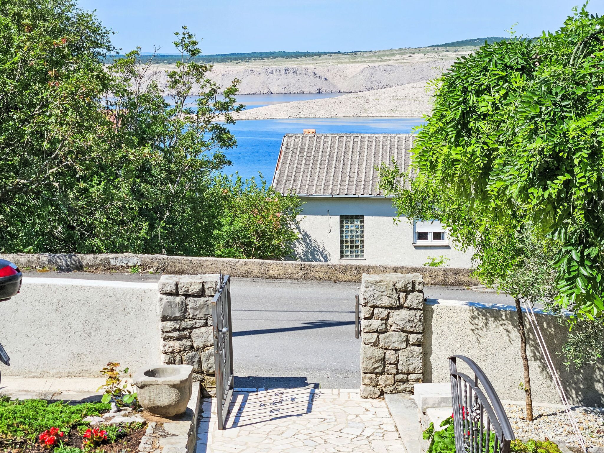 A stone gate leading to a paved path, with a view of a coastal landscape and a building with a tiled roof.