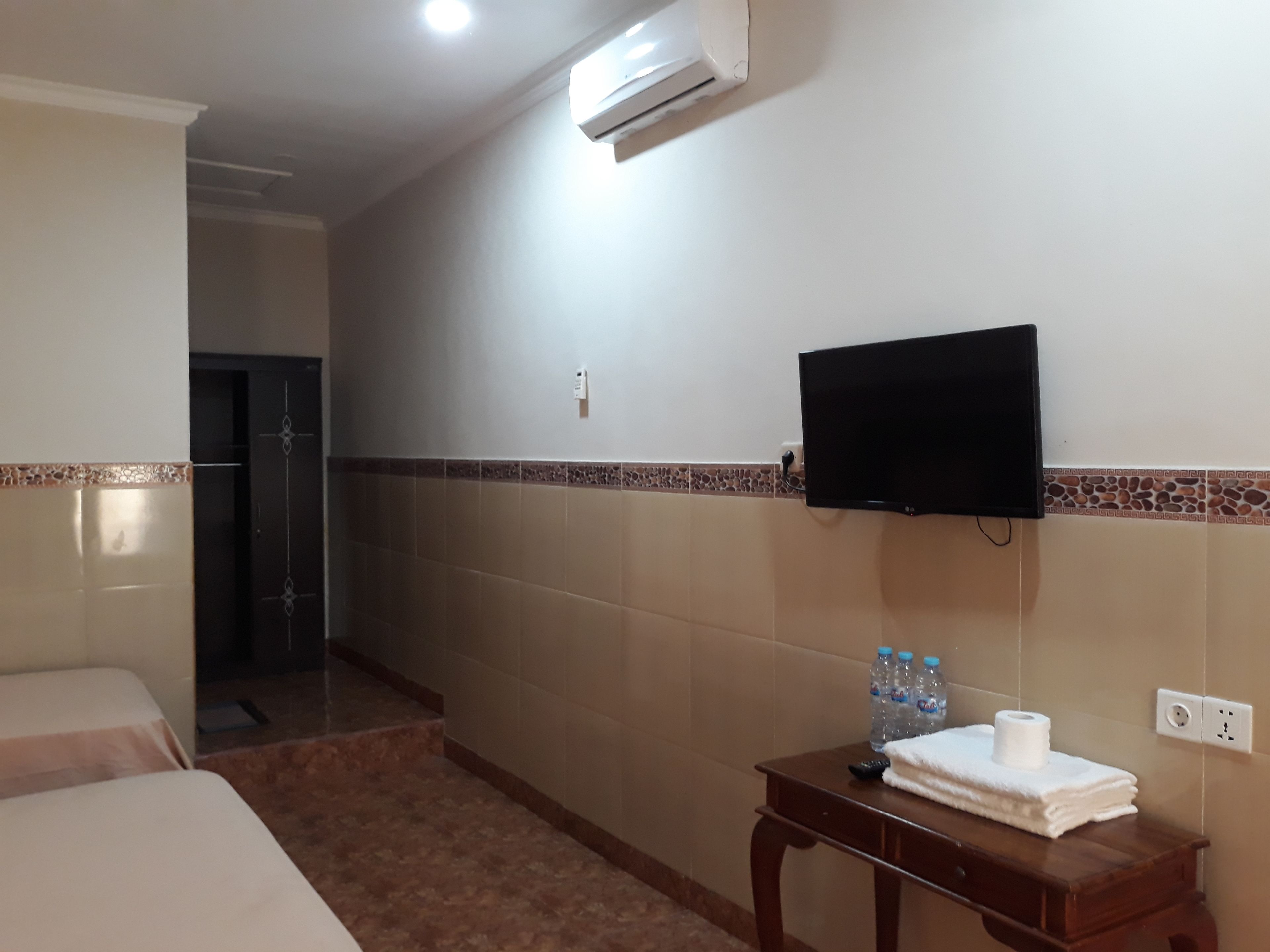 A hotel room with a bed, a small wooden table with toiletries, a wall-mounted TV, and an air conditioning unit.