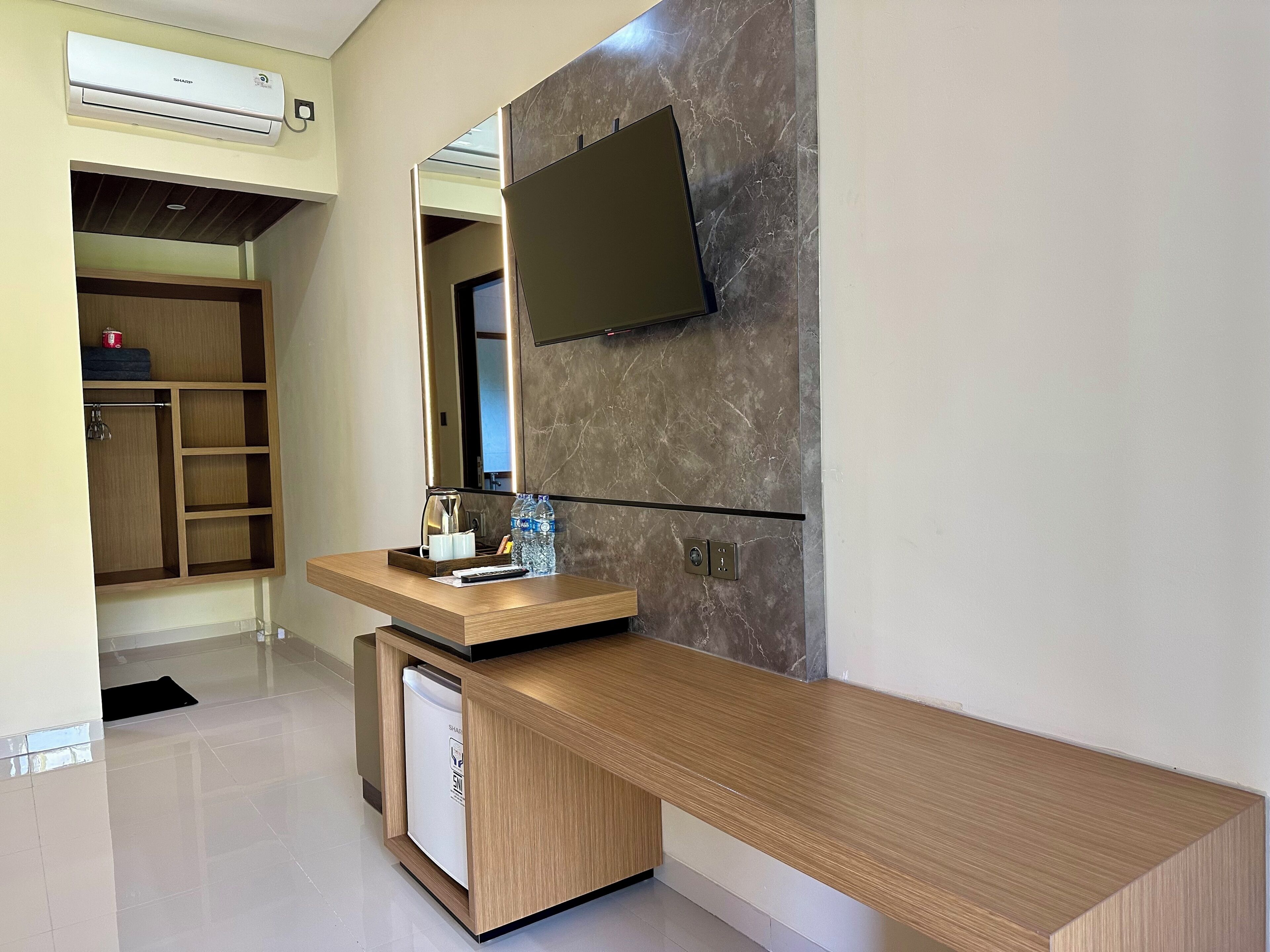 A modern hotel room with a wooden desk, a flat-screen TV mounted on the wall, a built-in trash bin, and a wardrobe with shelves.
