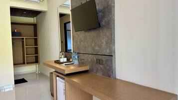A modern hotel room with a wooden desk, a flat-screen TV mounted on the wall, a built-in trash bin, and a wardrobe with shelves.
