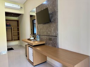 A modern hotel room with a wooden desk, a flat-screen TV mounted on the wall, a built-in trash bin, and a wardrobe with shelves.