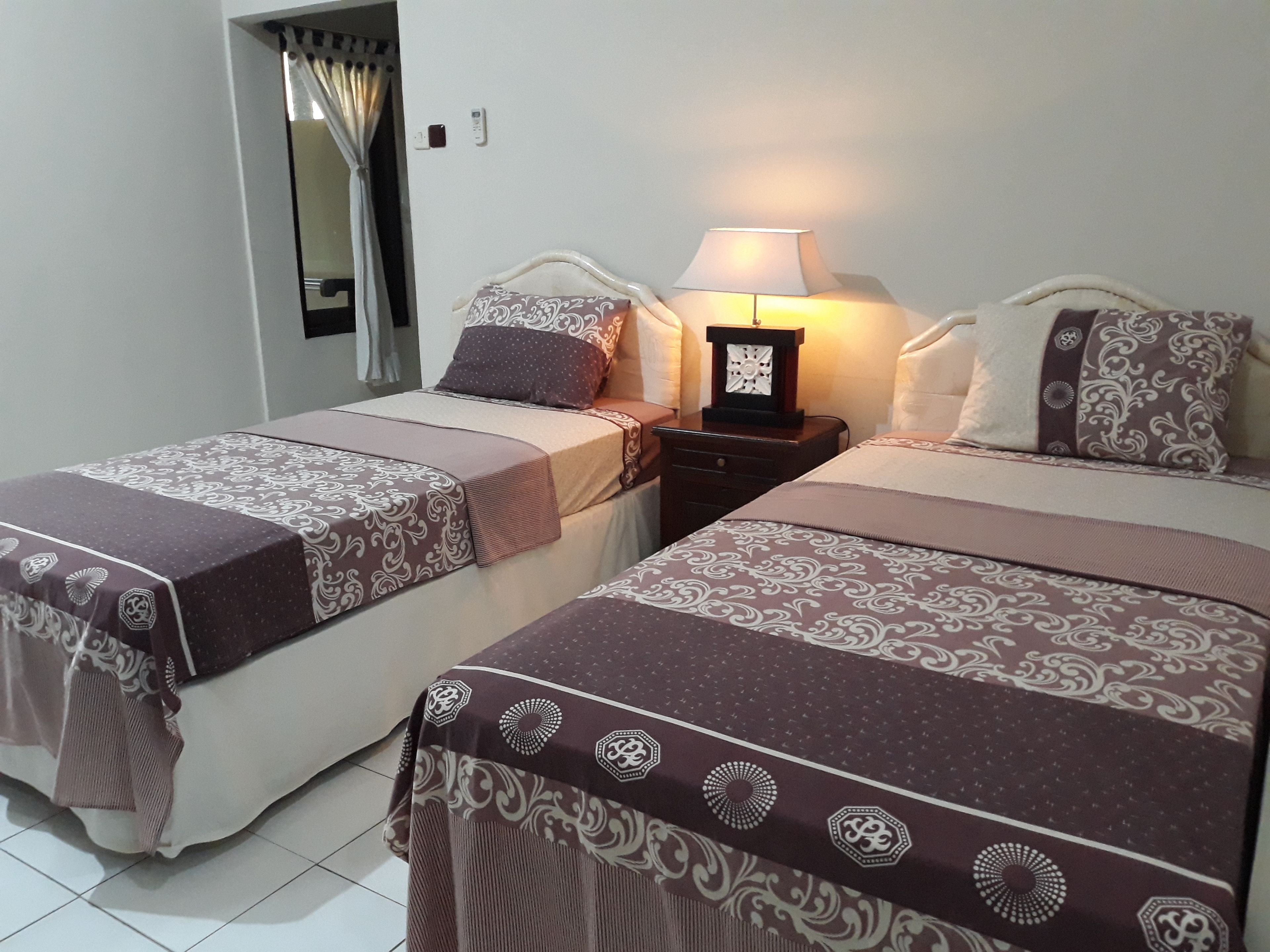 Una habitación de hotel con dos camas individuales, cada una con ropa de cama y almohadas estampadas, una mesita de noche con lámpara y un espejo en la pared.