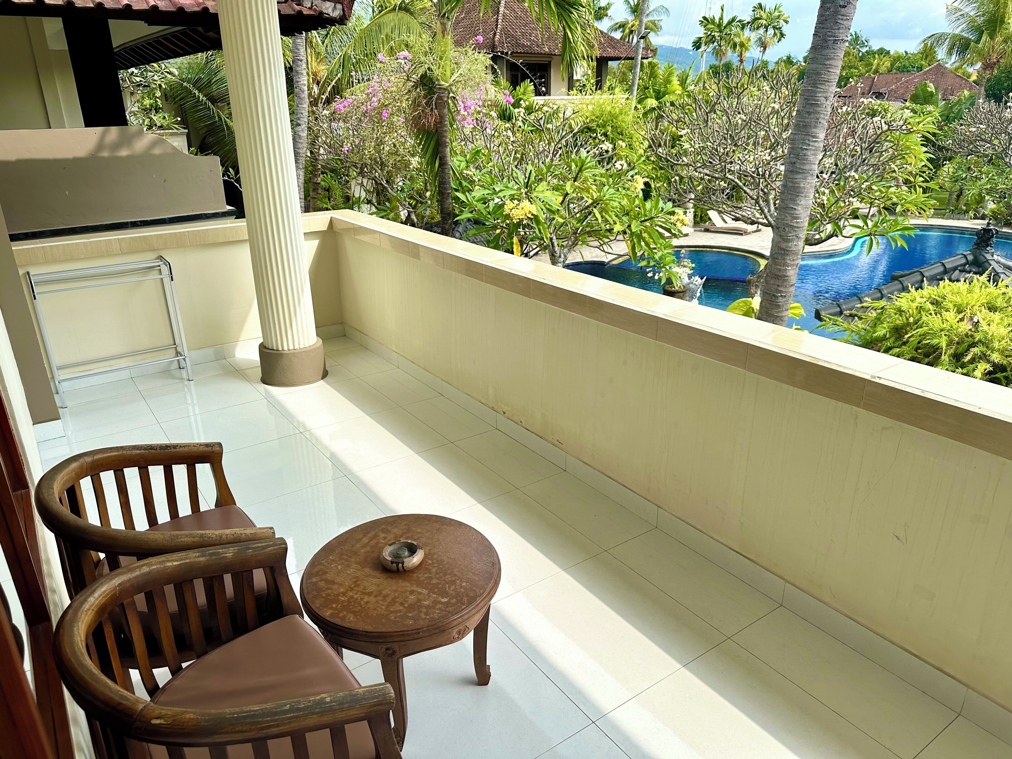 A balcony with a view of a pool, two wooden chairs, a small round table, and a tiled floor.