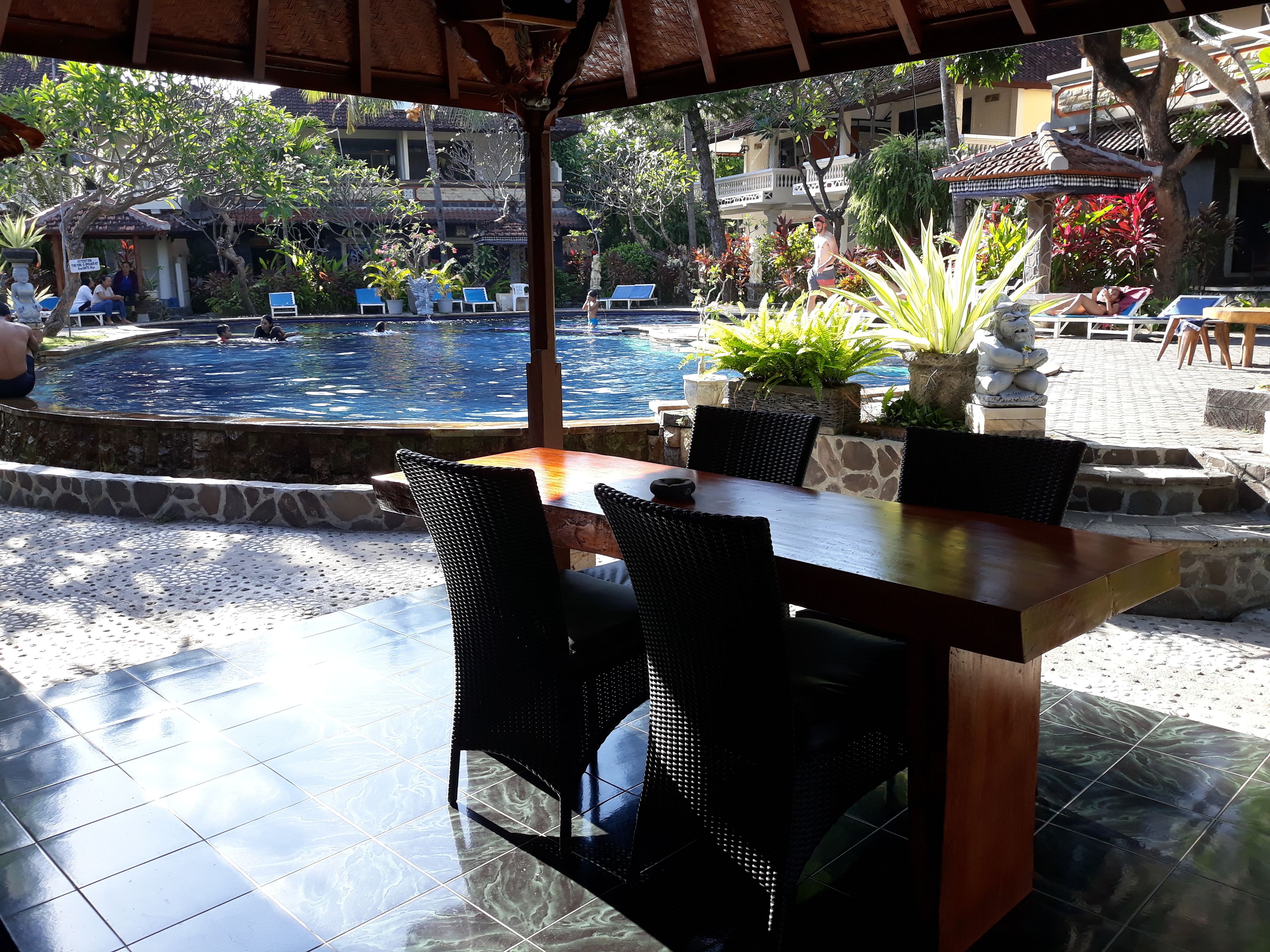 A poolside area with a wooden table and chairs, surrounded by potted plants and a statue.