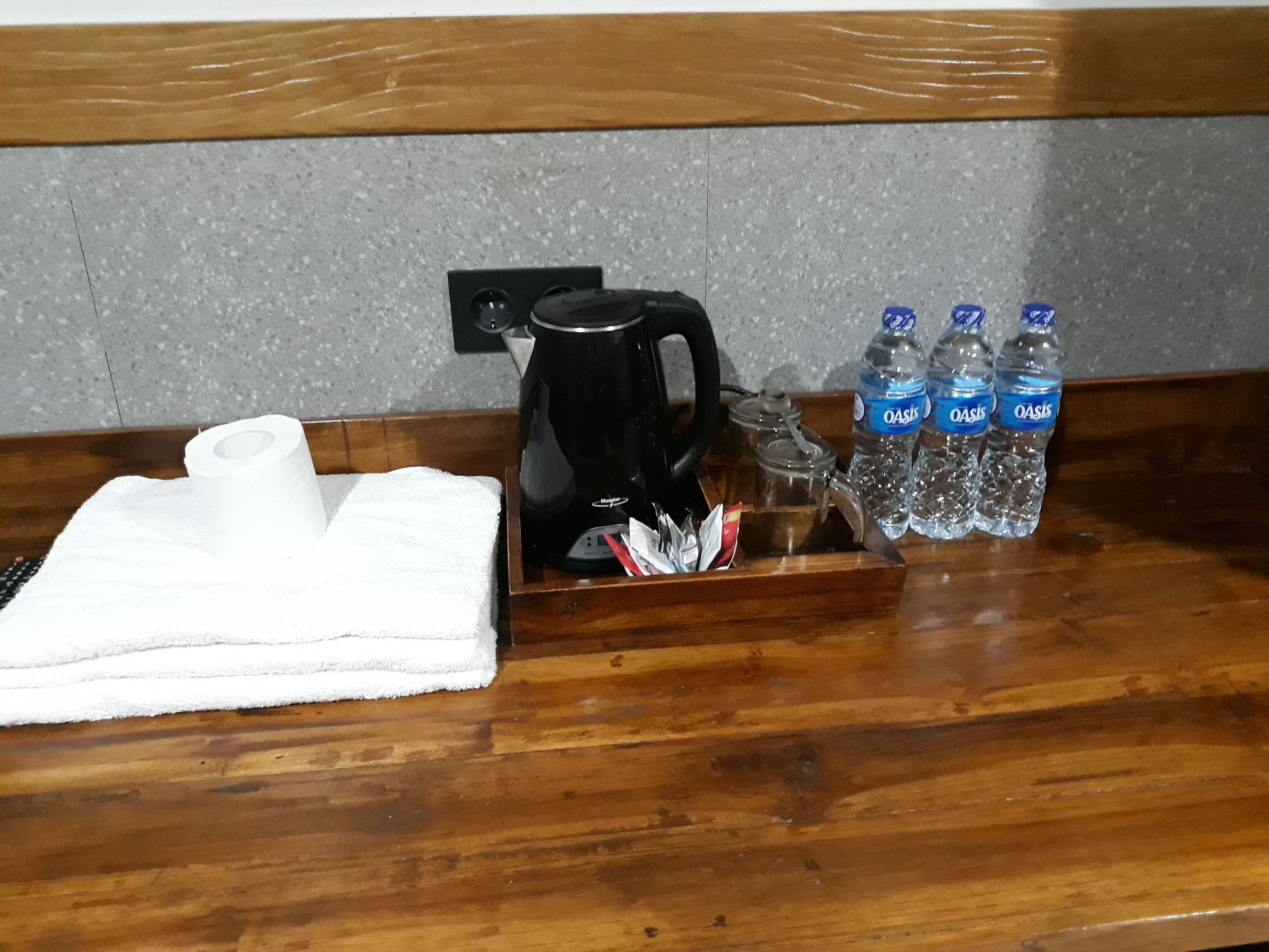A wooden counter with a black coffee maker, a roll of toilet paper, and three Oasis water bottles.