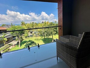 A balcony with a wicker chair, a view of a garden with palm trees, and a clear sky.
